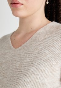 Beige knitted sweater with a V-neckline, featuring a textured, ribbed pattern and a soft, lightweight material.