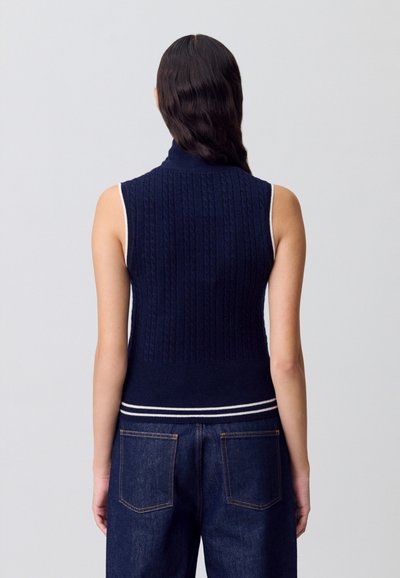 Suéter sin mangas de cuello alto en navy con textura de punto trenzado y ribete blanco a rayas, combinado con jeans de mezclilla oscura.