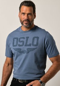 Modré bavlněné tričko s krátkými rukávy, s potiskem "OSLO" a grafikou křídel v tmavém provedení. Nosené s černými kalhotami a hodinkami.