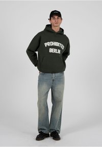 Bărbat purtând un hanorac verde închis cu textul „PROHIBITED BERLIN”, blugi deschiși la culoare, pantofi negri și o șapcă neagră, stând cu mâinile pe spate.