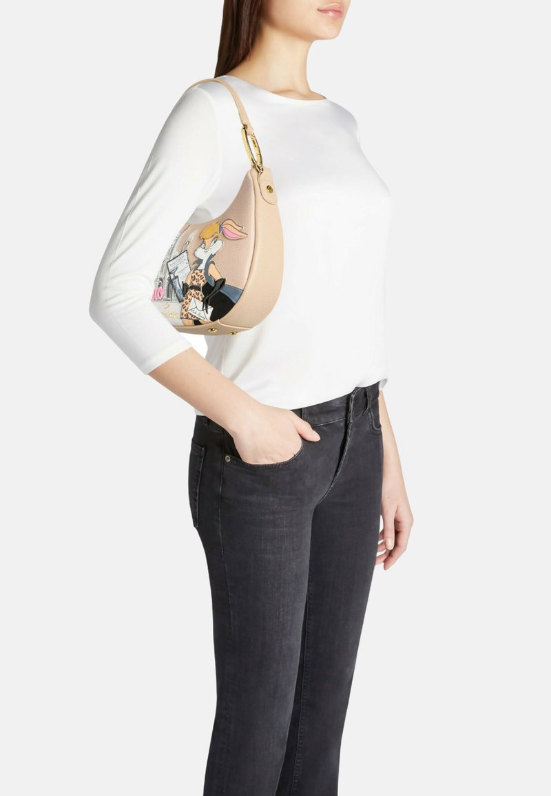 Borsa a tracolla in pelle color cuoio con grafica a fumetti, dettagli in oro e forma arrotondata. Indossata con una maglietta bianca e jeans neri.