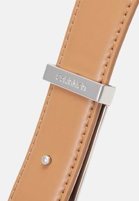 Calvin Klein kellosuojus ruskeasta nahasta, jossa on hopeanväriset yksityiskohdat, logo-kaiverrettu solki ja koristeellinen helmikoriste.