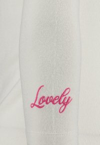 Giaccone bianco a maniche lunghe in maglia con ricamo rosa della scritta "Lovely" sul polsino, evidenziando una texture liscia e una forma aderente.