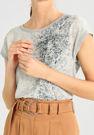 Vrouw draagt een grijs patroon shirt met korte mouwen, ingestopt in een beige broek met hoge taille, een stoffen riem en een ronde zilveren gesp.