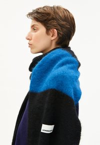 Profil d'une personne avec des cheveux bruns courts portant une écharpe floue noire et bleue avec une étiquette "ARMEDANGELS" sur fond blanc.