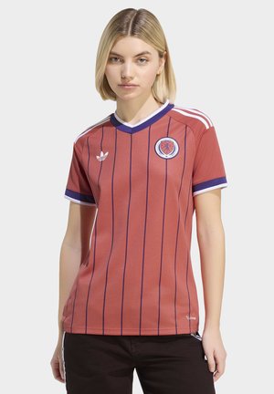 Donna bionda che indossa una maglia da calcio della Scozia a righe rosse con rifiniture blu navy e logo Adidas, in piedi davanti a uno sfondo semplice.