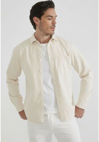 Homme portant une chemise beige à boutons par-dessus un t-shirt blanc et un pantalon blanc, se tenant contre un fond clair avec les mains sur les hanches.