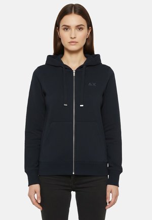 Giovane donna che indossa una felpa nera con zip semplice, con un piccolo logo sul petto a sinistra, e pantaloni neri, in piedi contro uno sfondo bianco.