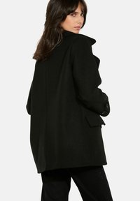 Schwarzer Wollmantel mit hohem Kragen, strukturierter Form, Knöpfen an den Ärmeln und Seitentaschen. Rückseite mit vertikaler Nahtdetail.