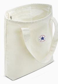 White canvas tote bag with two handles, featuring a circular red and blue logo on one side. Open top and interior zipper pocket.
