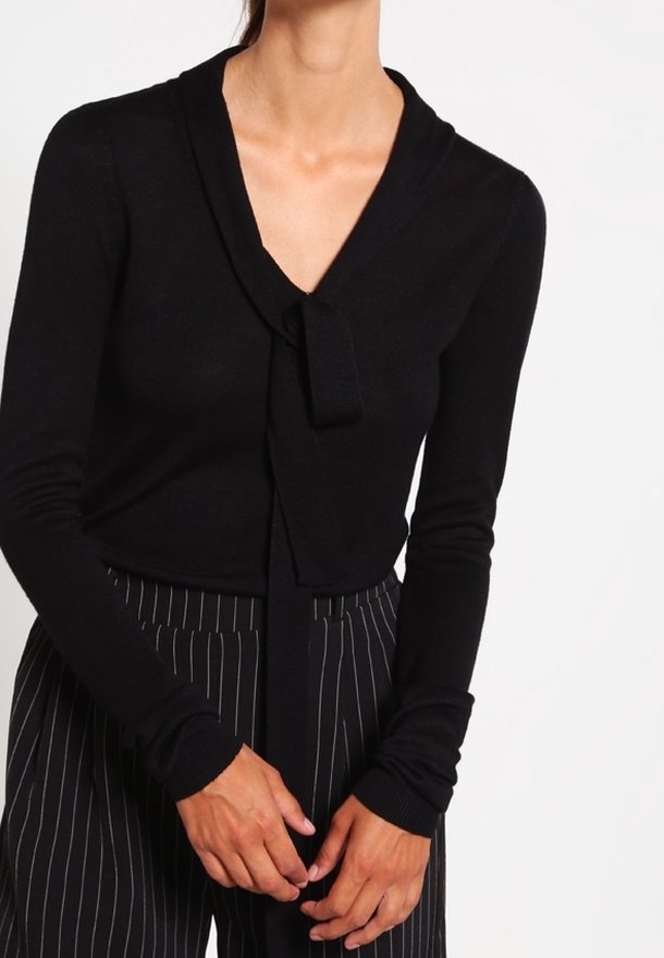 Personne portant un haut noir à manches longues avec un détail de nœud au col en V et un pantalon noir à fines rayures, les mains jointes devant.
