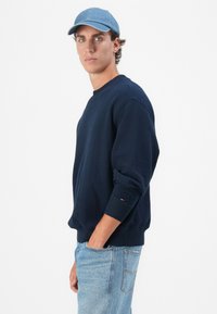 Navyblauwe sweater met textuur en ronde hals, met ribbels aan de mouwen, gecombineerd met lichtblauwe jeans. Een dennenpet wordt op het hoofd gedragen.