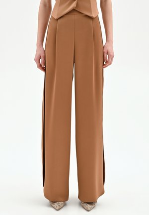 Pantaloni marroni a gamba larga con pieghe frontali, spacchi laterali, indossati con tacchi beige a punta su uno sfondo bianco.