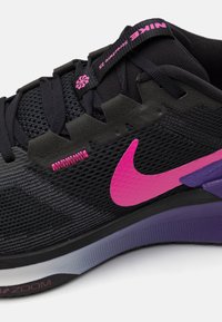 Crni Nike tenisice s mrežastim gornjim dijelom, ružičastim naglascima i ljubičastim potplatom; sadrže jedinstveni dizajn vezica i vidljive elemente amortizacije.