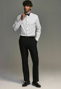 Camisa blanca con botones negros y cuello alzado, combinada con pantalones negros y zapatos negros brillantes, acentuada con una pajarita negra.