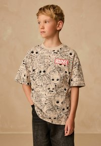 Beige kortærmet t-shirt med sorte stregtegninger af Marvel-figurer og et rødt "MARVEL" logo-badge på brystet.