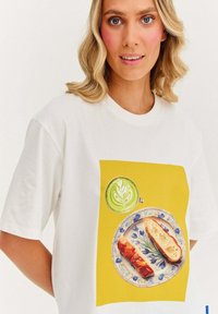 Bílý tričko s grafikou žlutého pozadí, na kterém je talíř s dvěma jídly a zelené latte art v hrnku.