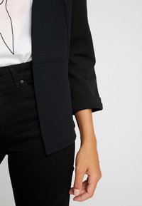 Blazer noir à texture lisse, avec manches retroussées et une poche avant. Associé à un jean foncé et un T-shirt blanc avec un motif graphique.