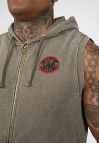Hoodie zonder mouwen in olijfgroen, met een frontale rits en een cirkelvormig rood 'Gorilla Wear' logo met een zwart gorilla-illustratie.