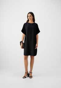 Svart kortärmad klänning med en lös passform, rund halsringning och strukturerad tyg. Matchad med en liten svart clutch och svarta klackskor med remmar.