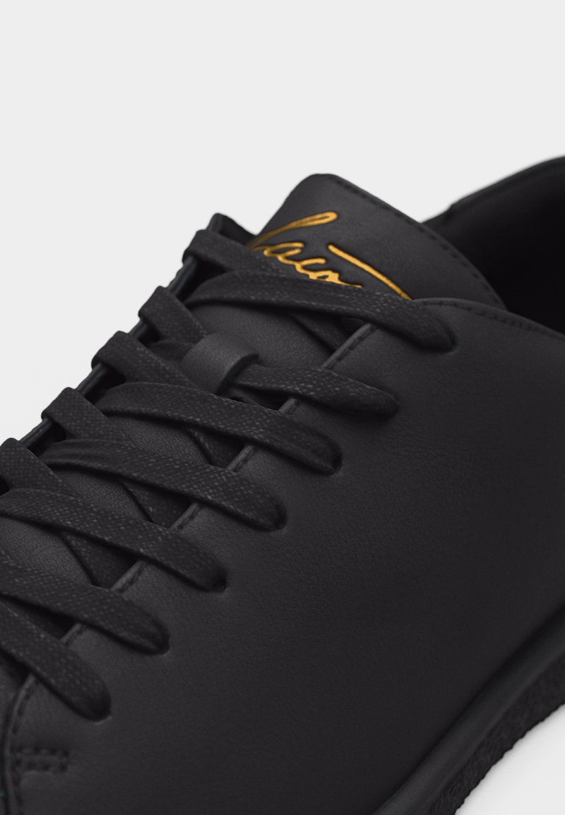 Zwarte leren sneaker met zwarte veters en gouden scriptlogo op de tong, in een close-up vanuit een schuine bovenaanzicht.