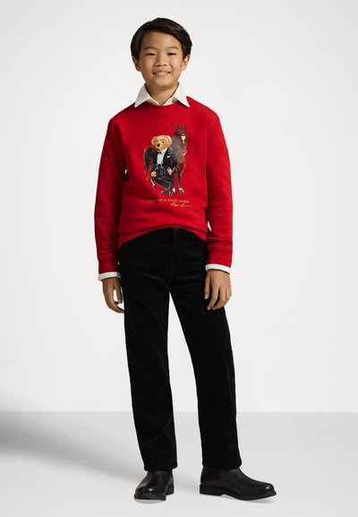 Sweatshirt vermelho com uma estampa de urso e cavalo, combinado com calças de bombazine pretas e botas pretas. Camisa branca com gola visível nas mangas.