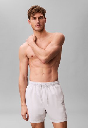 Homme torse nu aux cheveux courts et bouclés, portant un short blanc à cordon, debout devant un fond gris uni, tenant son épaule gauche.