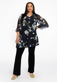 Zwarte tuniek met bloemmotieven in blauw en groen, voorzien van doorzichtige belmouwen. Gepaard met zwarte broek en groene haksandalen.