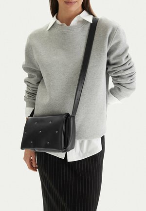 Žena v šedém svetru a bílé košili nese černou koženou crossbody kabelku s cvočky a dlouhým popruhem.