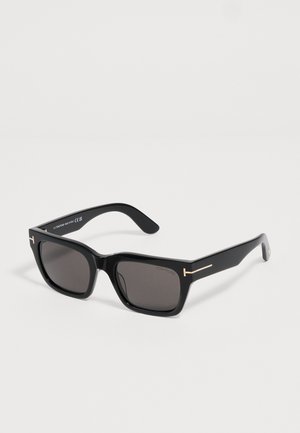 Schwarze, rechteckige Tom Ford Sonnenbrille mit dunklen Gläsern und goldenen T-förmigen Akzenten an den Bügeln, liegend auf einer weißen Oberfläche.