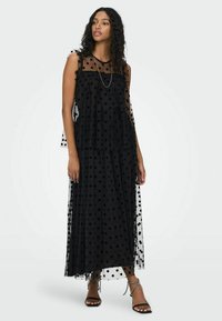 Maxi dress nera a pois bianchi in tessuto trasparente con design a strati, caratterizzata da maniche a volant e una silhouette fluttuante, indossata con sandali neri con cinturino.