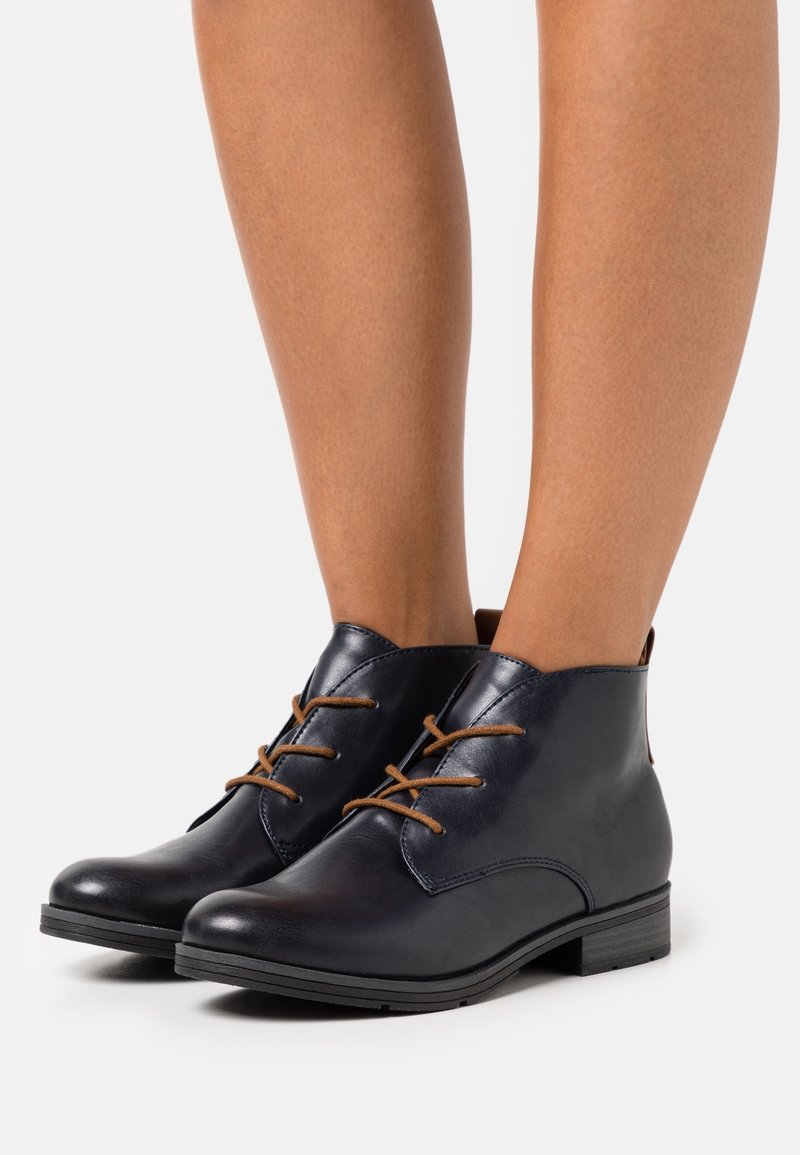 Bottines en cuir noir à lacets marron portées sur des jambes nues sur un fond blanc uni.