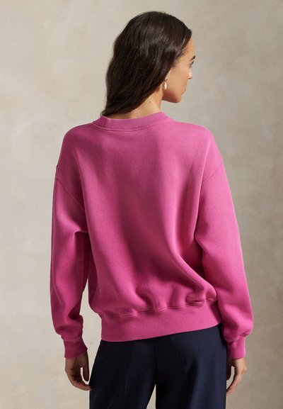 Γυναίκα με σκούρα μαλλιά που φορεί φωτεινό ροζ φούτερ με μακριά μανίκια και σκουρόχρωμο παντελόνι, στέκεται με την πλάτη προς έναν μπεζ τοίχο.