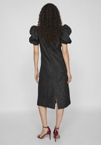 Μαύρο φόρεμα με υφή υφάσματος, φουσκωτά μανίκια, μήκος μέχρι το γόνατο και σχισμή στο πίσω μέρος. Φορεμένο με κόκκινες ψηλοτάκουνες σαγιονάρες.