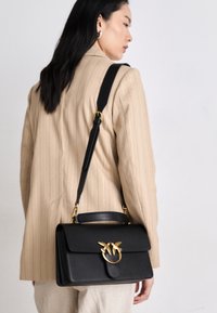 Čierna kožená kabelka so štruktúrovaným dizajnom, zlatými akcentmi a dekoratívnym uzáverom s motívom motýľa. Nastaviteľný ramenný popruh.