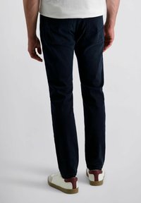 Mann trägt dunkelblaue Jeans und weiße Sneakers mit braunen Akzenten, steht vor einem schlichten hellen Hintergrund.