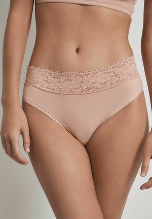 Femeie care poartă lenjerie intimă nude cu talie înaltă și dantelă florală, stând cu mâinile relaxate pe lângă corp pe un fundal simplu.