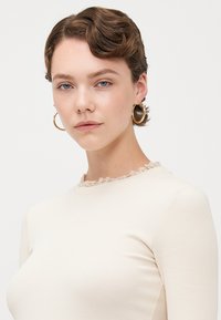Haut en maille côtelée beige clair avec un col rond, orné d'une fine bordure froncée. Modèle portant des boucles d'oreilles en or en forme de hoop, texture lisse.