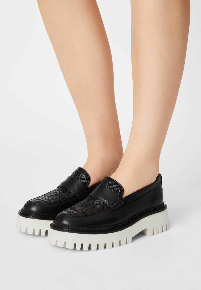 Mocassins en cuir noir avec une tige texturée et de épais semelles à crampons blanches portées pieds nus sur un fond blanc.