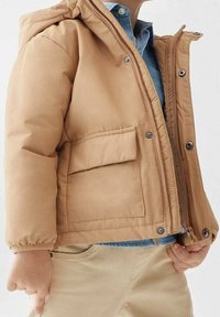 Giacca puffer beige con chiusura a zip e bottoni a pressione, cappuccio e tasca frontale. Presenta polsini elasticizzati e una texture liscia e opaca.