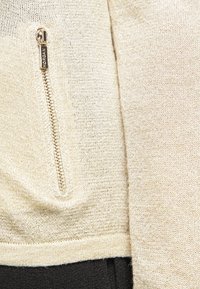 Stickad plagg i ljusbeige tyg med en subtil lyster, med en synlig gulddragkedja på vänster sida. Strukturerad yta med en slät finish.