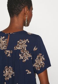 Vestido marinho de mangas curtas, com um padrão floral em branco e dourado. Detalhe nas costas em forma de olho de fechadura acrescenta um toque decorativo. Textura suave.