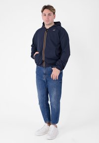 Giacca a vento blu con cappuccio, zip gialla, polsini elastici e due tasche frontali, abbinata a jeans in denim azzurro chiaro e sneakers bianche.