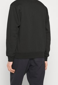 Felpa nera con collo rotondo e polsini a coste, caratterizzata da una texture morbida. Indossata con pantaloni blu scuro, vista da dietro.