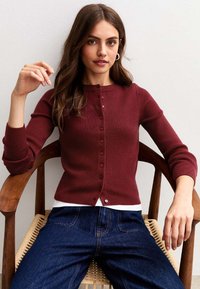 Bordeaux ribbeltrui met een ronde hals en vijf knopen, gecombineerd met een witte blouse en blauwe spijkerbroek, zittend op een houten stoel.