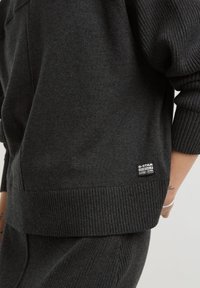 Pull en maille côtelée gris foncé et pantalon avec l'étiquette G-Star Originals Raw Denim sur le pull, montrant une partie d'un bras et d'une main tatoués.