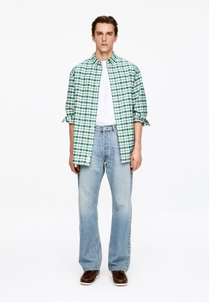 Koszula w zielono-białą kratę na guziki, nałożona na białą koszulkę, do tego jasnoniebieskie dżinsy i brązowe buty. Prosty wzór i kolory.