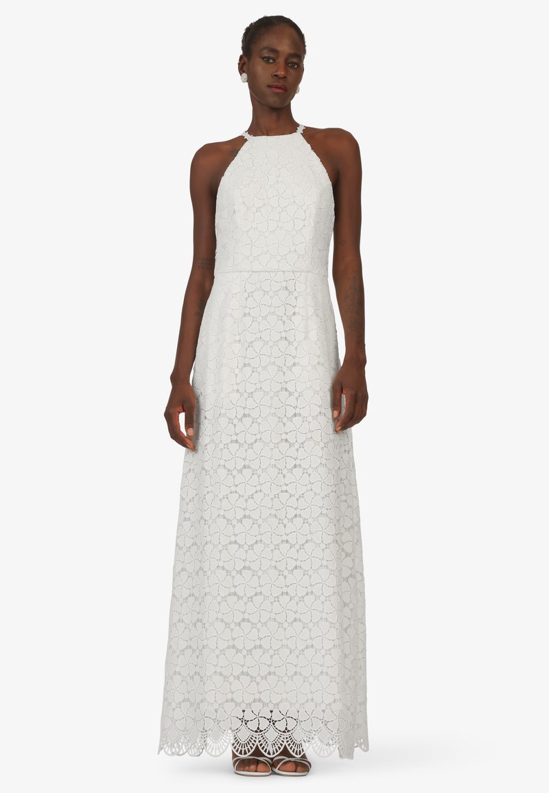 Witte kanten maxi-jurk met halternek, bloemenpatroon en golvende rand. Zachte textuur en verlengde silhouet, gecombineerd met open teen schoenen.
