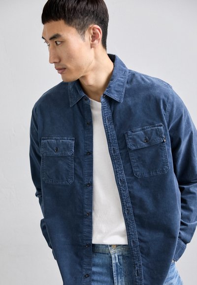 Camisa de algodón azul con dos bolsillos en el pecho, cierre de botones en la parte delantera y cuello puntiagudo. Lleva puesta sobre una camiseta blanca, combinada con jeans de mezclilla.
