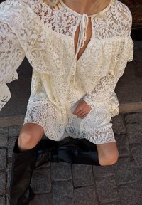 Weißes Spitzenkleid mit gerüschten Lagen und einer Schleifen-Kragen. Schwarze kniehohe Stiefel ergänzen das Outfit. Strukturierte Spitzen-Details sind auffällig.